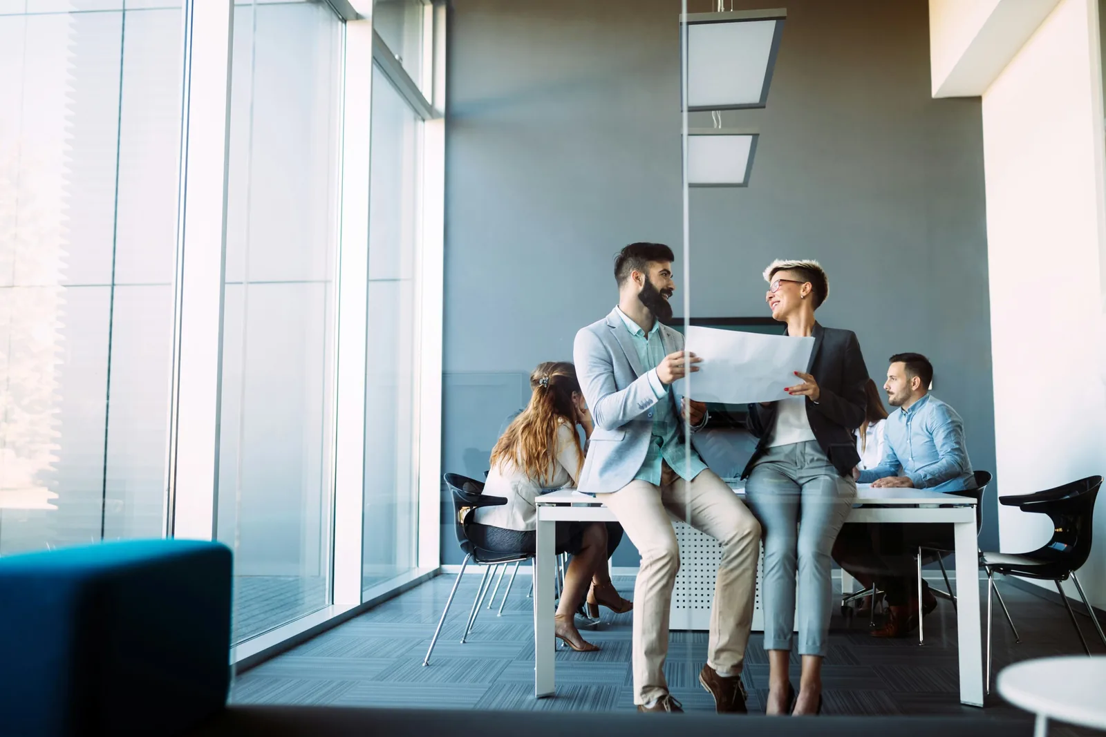 Zwei Kollegen besprechen Dokumente in einem modernen Büro mit großen Fenstern und weiteren Mitarbeitern im Hintergrund.